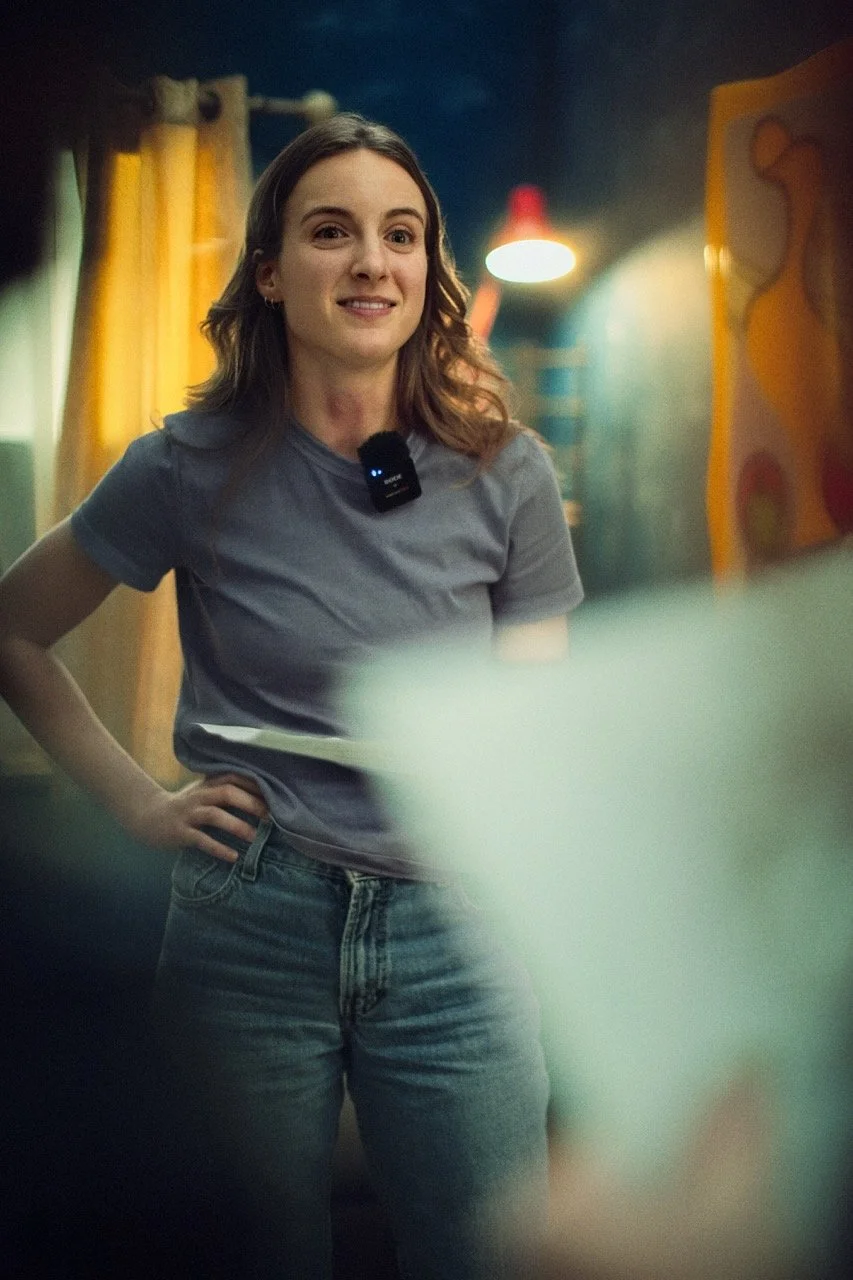 A young woman with brown wavy hair, wearing a gray t-shirt and jeans, stands with one hand on her hip in a room with colorful decor and a red hanging lamp.