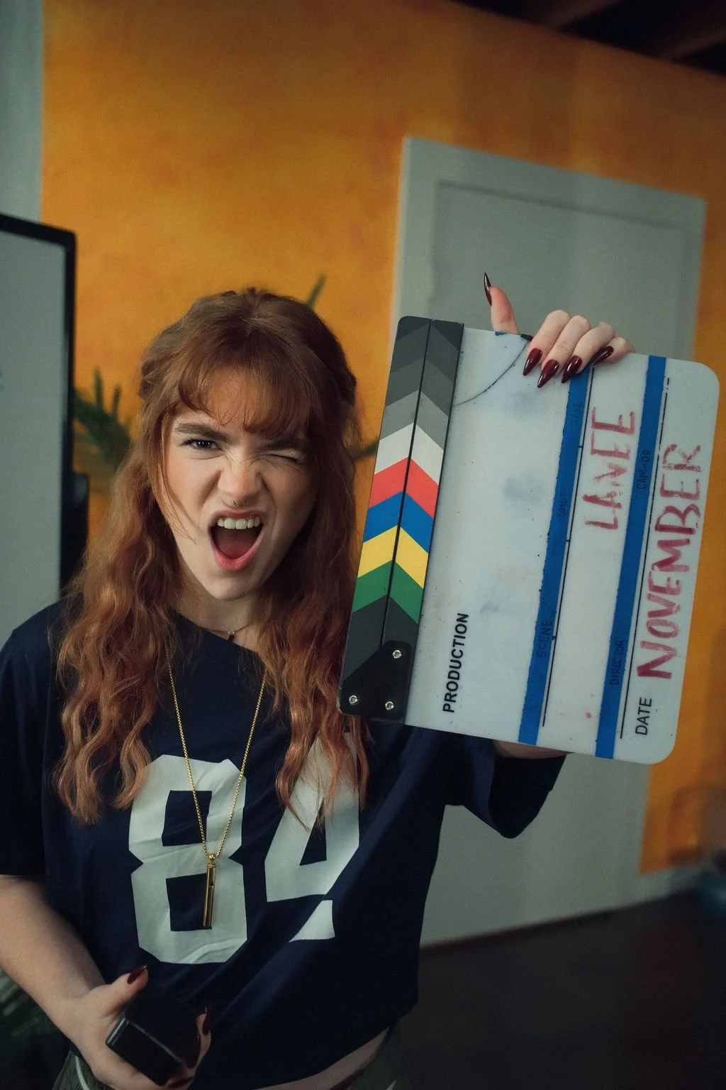 A young woman with red hair making a playful face and winking, holding a film slate with the words 'LAINE NOMEMBER' written on it, in an indoor setting with an orange wall in the background.
