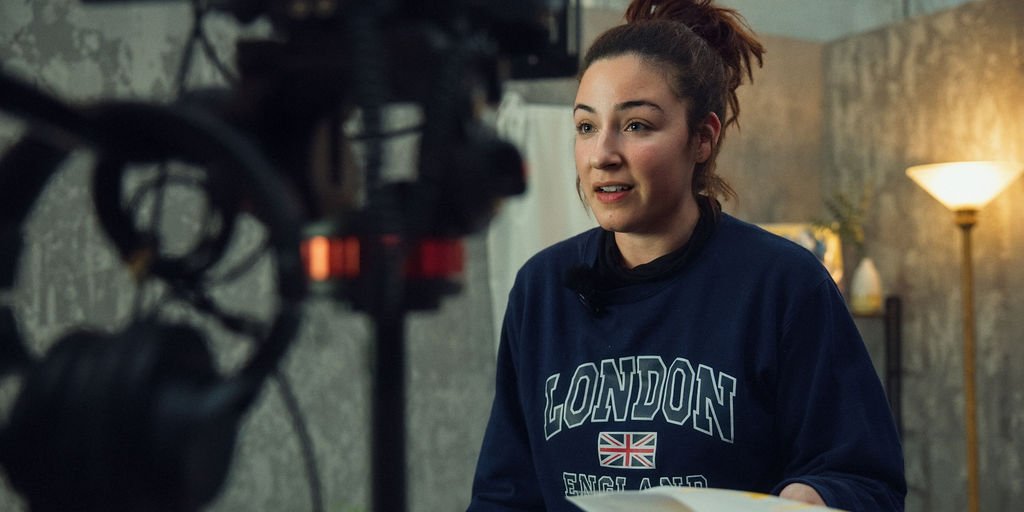 A woman with a bun hairstyle speaking in front of a camera, wearing a navy sweatshirt with 'LONDON, ENGLAND' printed on it, in a cozy indoor setting with a lamp and a textured wall.