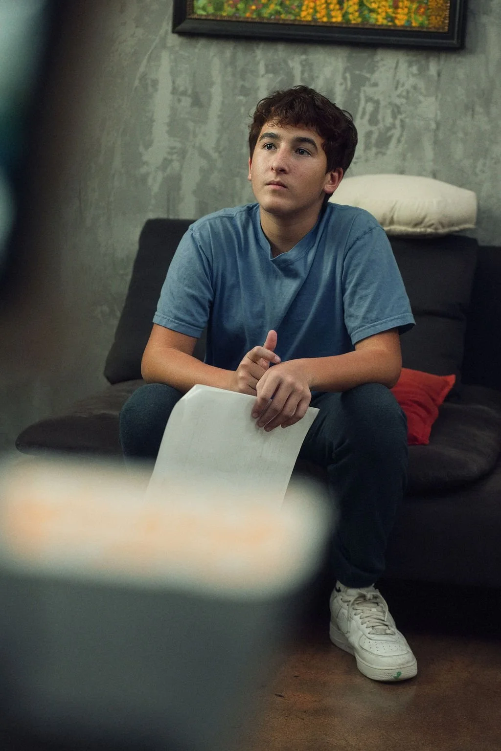 A teenage boy sitting on a dark sofa, holding a sheet of paper, with a contemplative expression in a room with gray textured walls and a colorful painting hanging above him.
