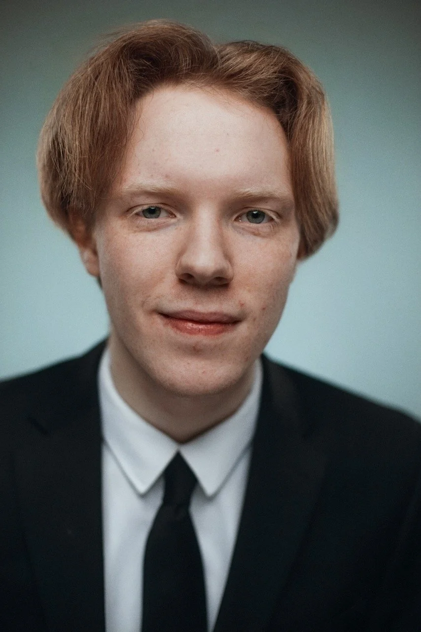 A young man with reddish-brown hair, fair skin, wearing a black suit, white shirt, and black tie, against a plain background.