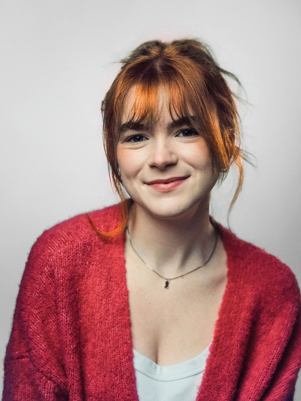 Young woman with red hair, wearing a white headband and a gray graphic t-shirt, smiling while holding a camera in her right hand against an orange background.