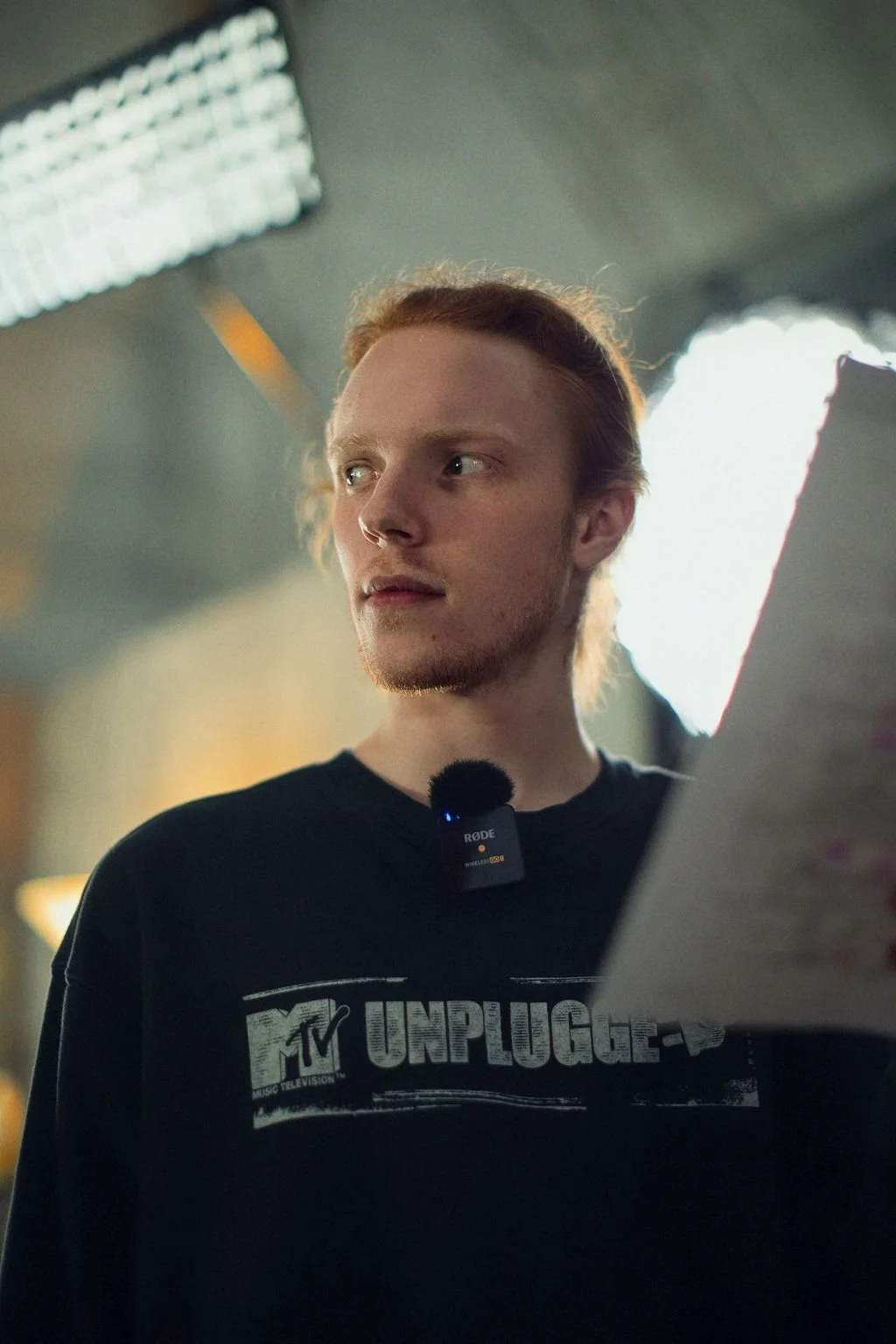 A young man with red hair and a beard wearing a black MTV Unplugged t-shirt, with a microphone clipped to his shirt, looking to the side in a studio with bright lights.