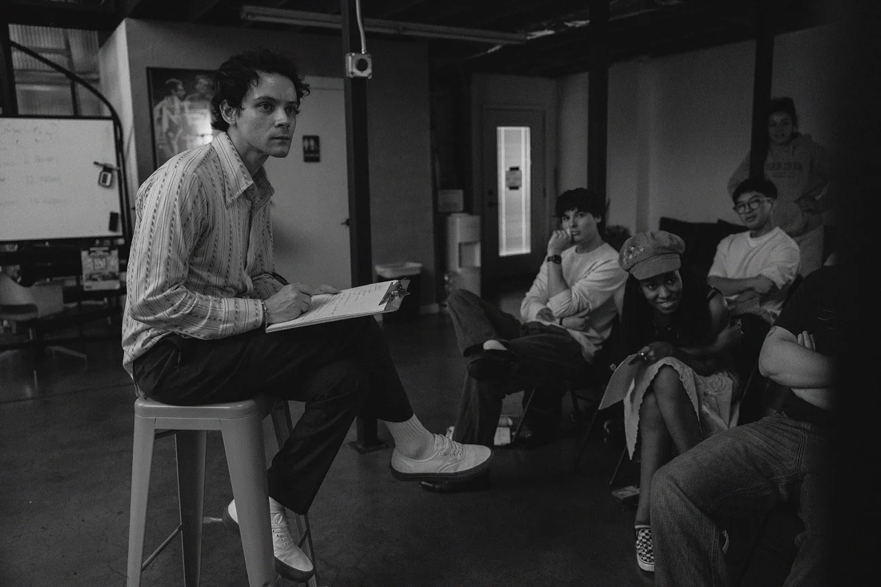 A young man sitting on a stool, holding a clipboard, appears to be speaking or presenting to a group of five people seated and listening intently in a casual indoor setting.