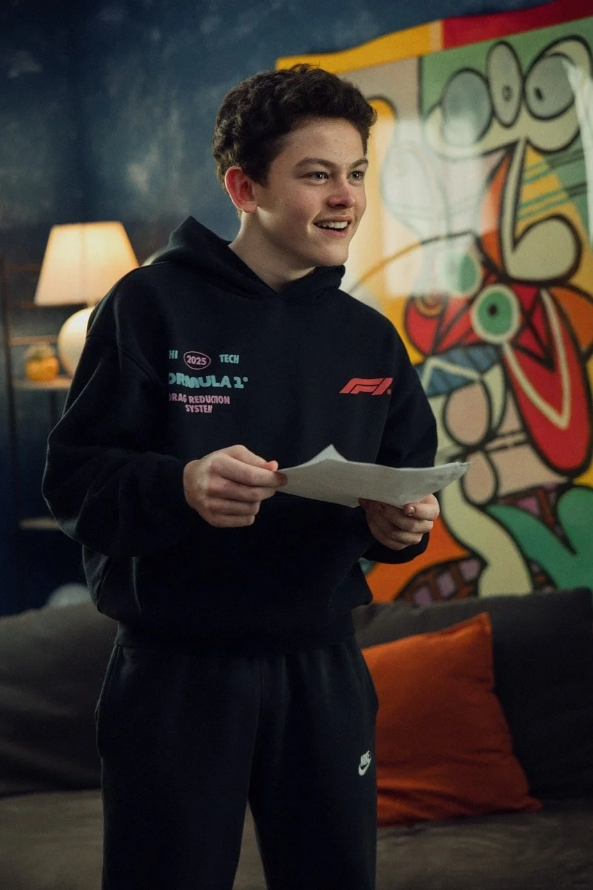 A young man with curly hair smiling and holding a paper, wearing a black hoodie and black Nike pants, standing in a room with a colorful graffiti-style painting and a lamp in the background.