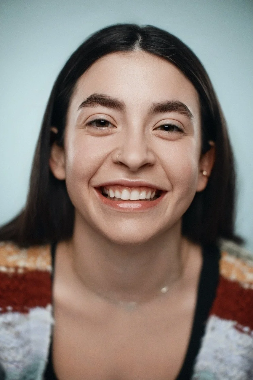 Close-up of a young woman smiling with missing teeth, wearing a colorful sweater and small jewelry, against a neutral background.