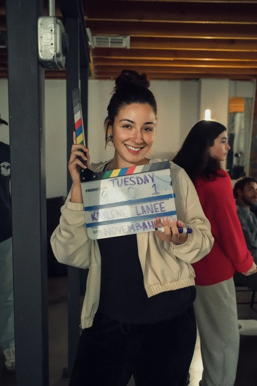 Young woman smiling and holding a clapperboard with information about a film or video production, surrounded by people in an indoor setting.
