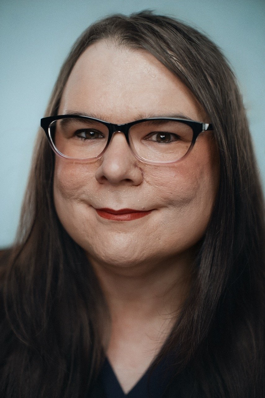 Close-up portrait of a woman with long brown hair, wearing black glasses, and smiling softly. She has light skin and is dressed in a dark top against a light background.