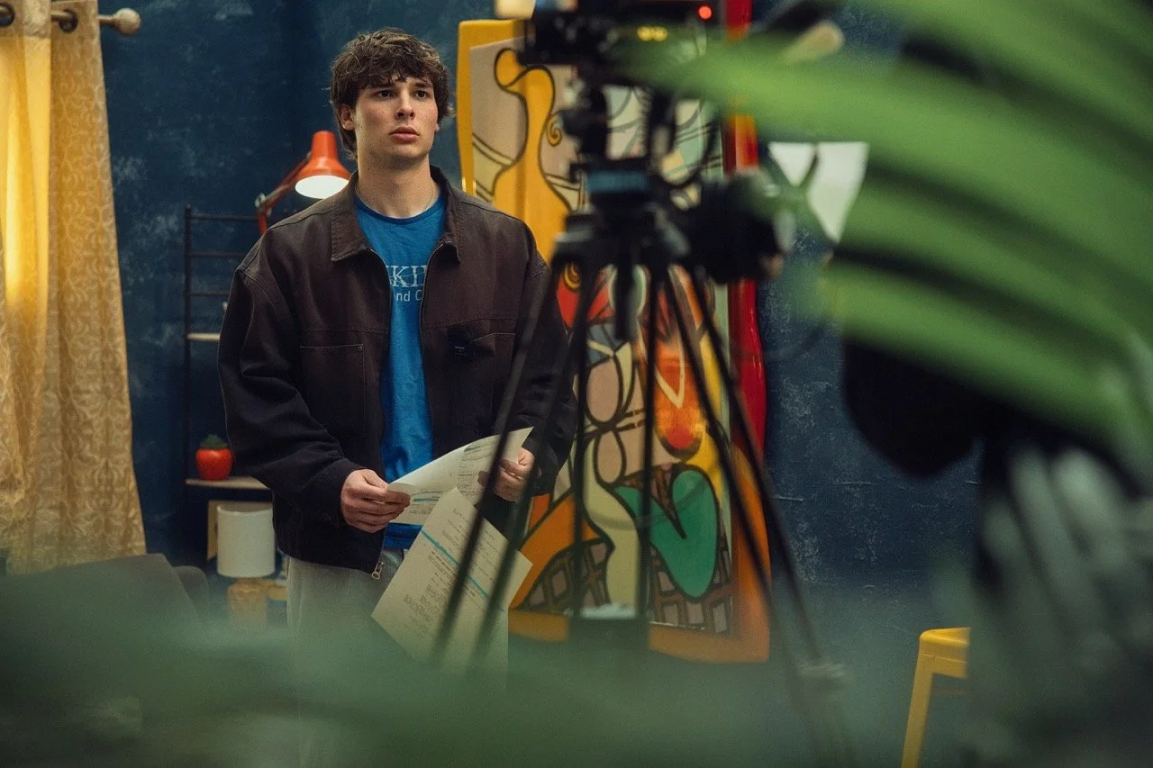 A young man with brown curly hair in a dark jacket holding papers, standing in front of a camera on a tripod in an indoor setting with colorful artwork and decor.