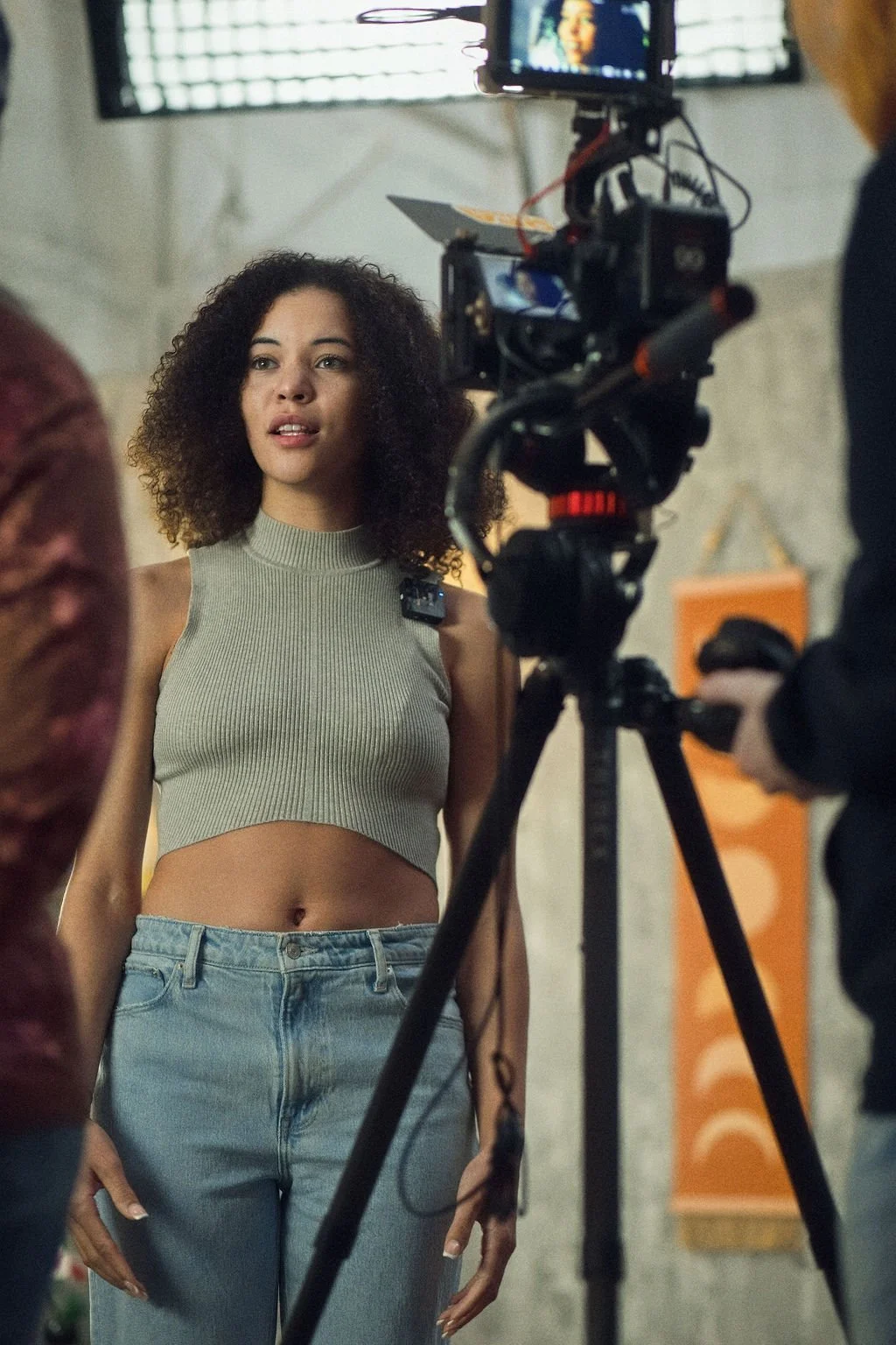 A young woman with curly hair is filming or being filmed on a camera in a studio. She is wearing a sleeveless, ribbed gray crop top and high-waisted jeans.