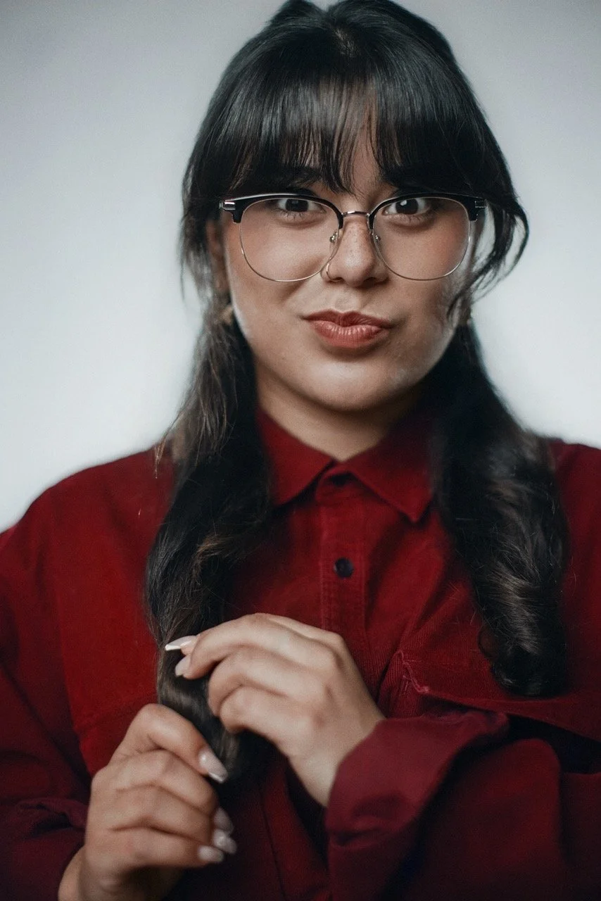 A woman with long dark hair and glasses, wearing a red shirt, is holding her hair and looking at the camera.