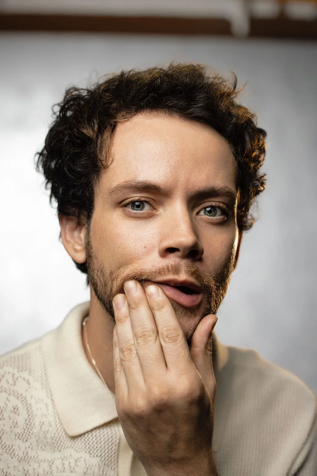 Portrait of a young man with curly dark hair and fair skin, wearing a cream short-sleeved shirt with a patterned design, standing in front of a vibrant orange and pink gradient background.