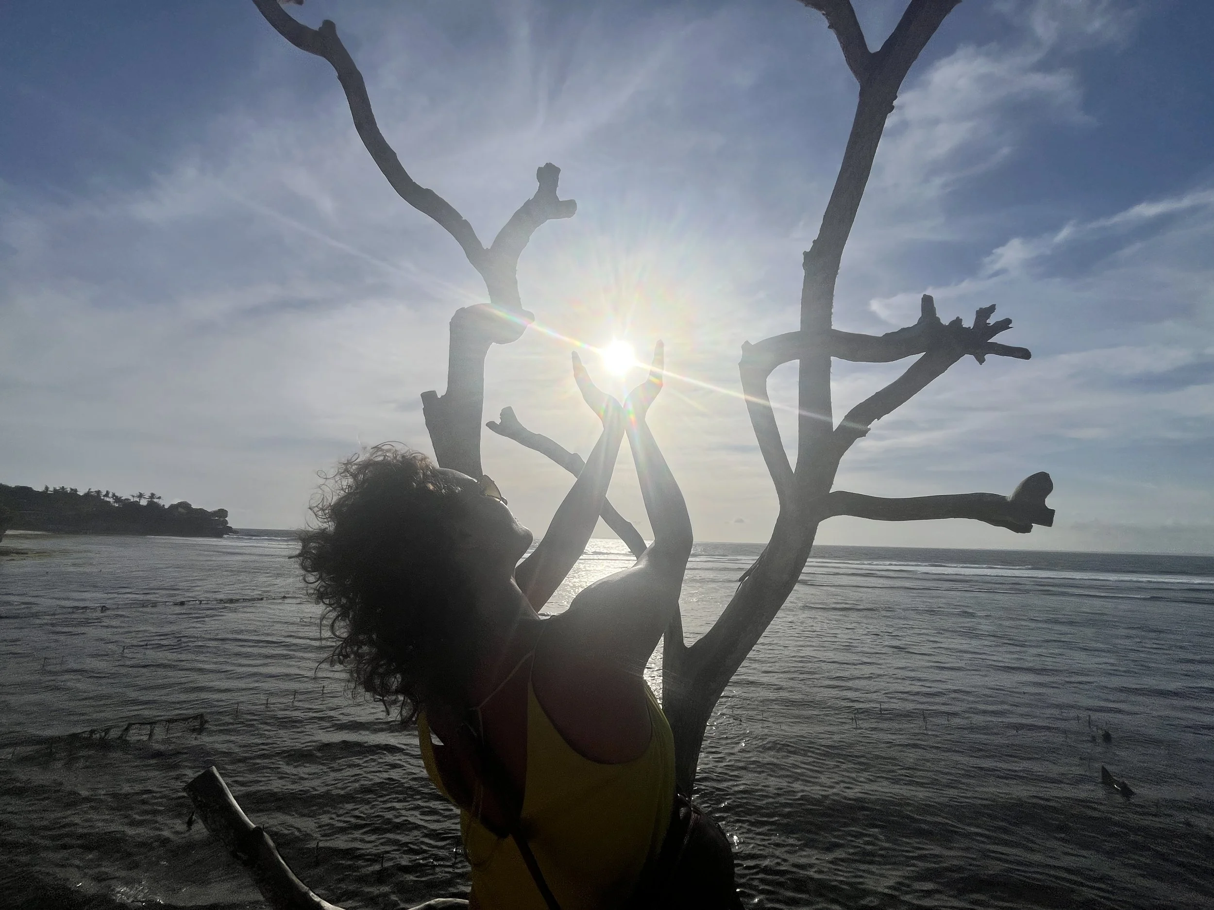 Eveline staat aan zee met haar gezicht naar de zon en haar armen geopend naar het licht, in een moment van verbinding, levensenergie en aanwezigheid.