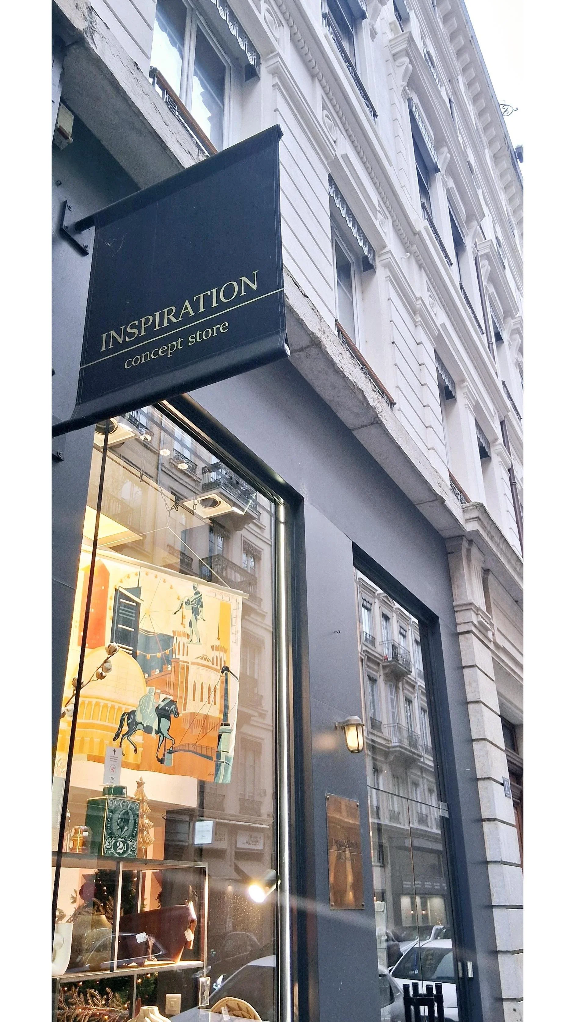 Façade extérieure d'un magasin nommé "INSPIRATION concept store" avec une vitrine affichant des objets décoratifs et une illustration suspendue avec un cheval et une architecture monumentale.