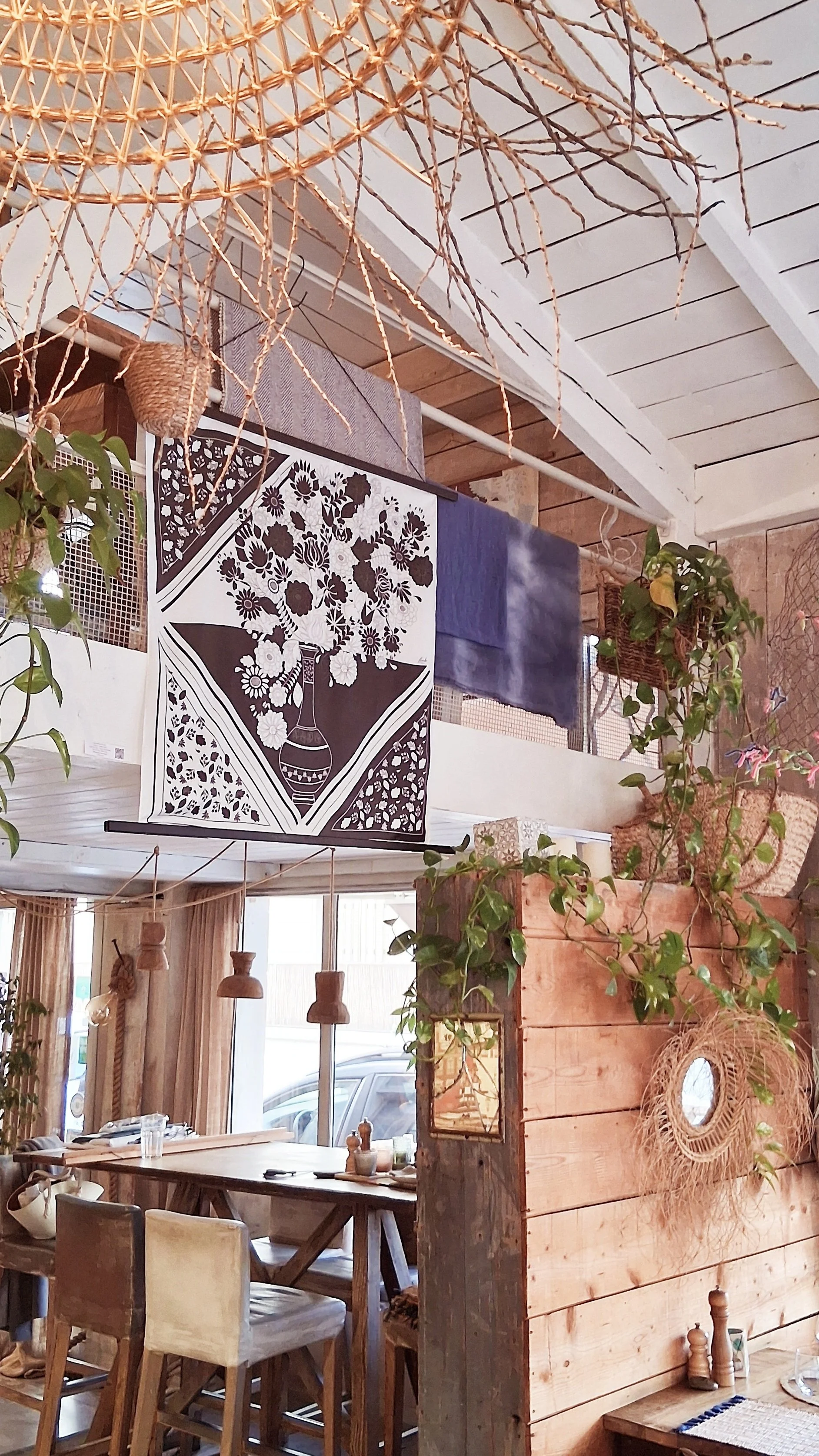 Intérieur d'un restaurant avec décor en bois, plantes suspendues, une affiche noire et blanche avec un bouquet de fleurs en vase, et un mur en bois avec miroir.