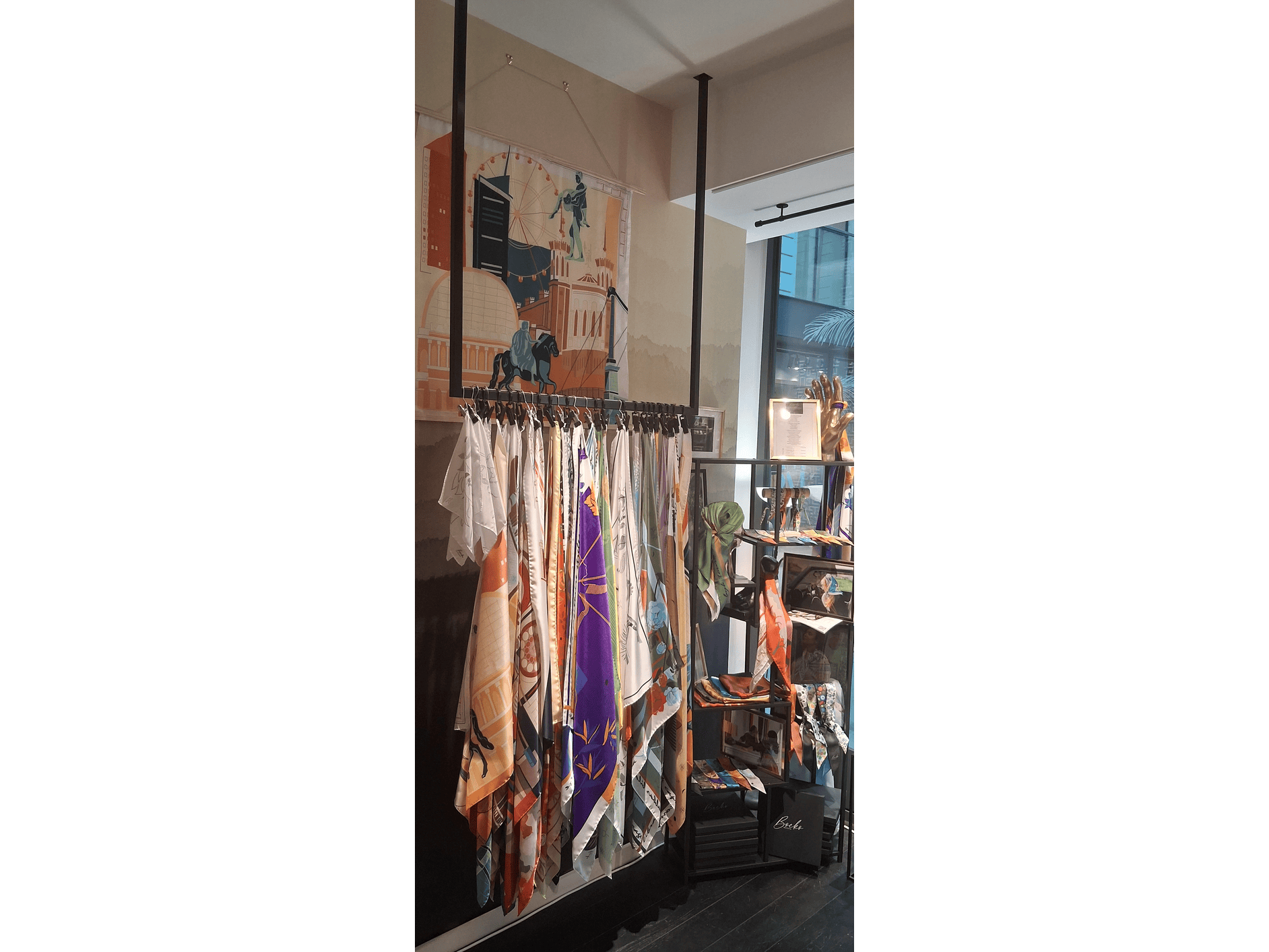 Vêtements et accessoires en vente dans une boutique, avec des écharpes colorées suspendues sur un support en métal et des lampes lumineuses, dans un décor moderne avec une grande fenêtre.