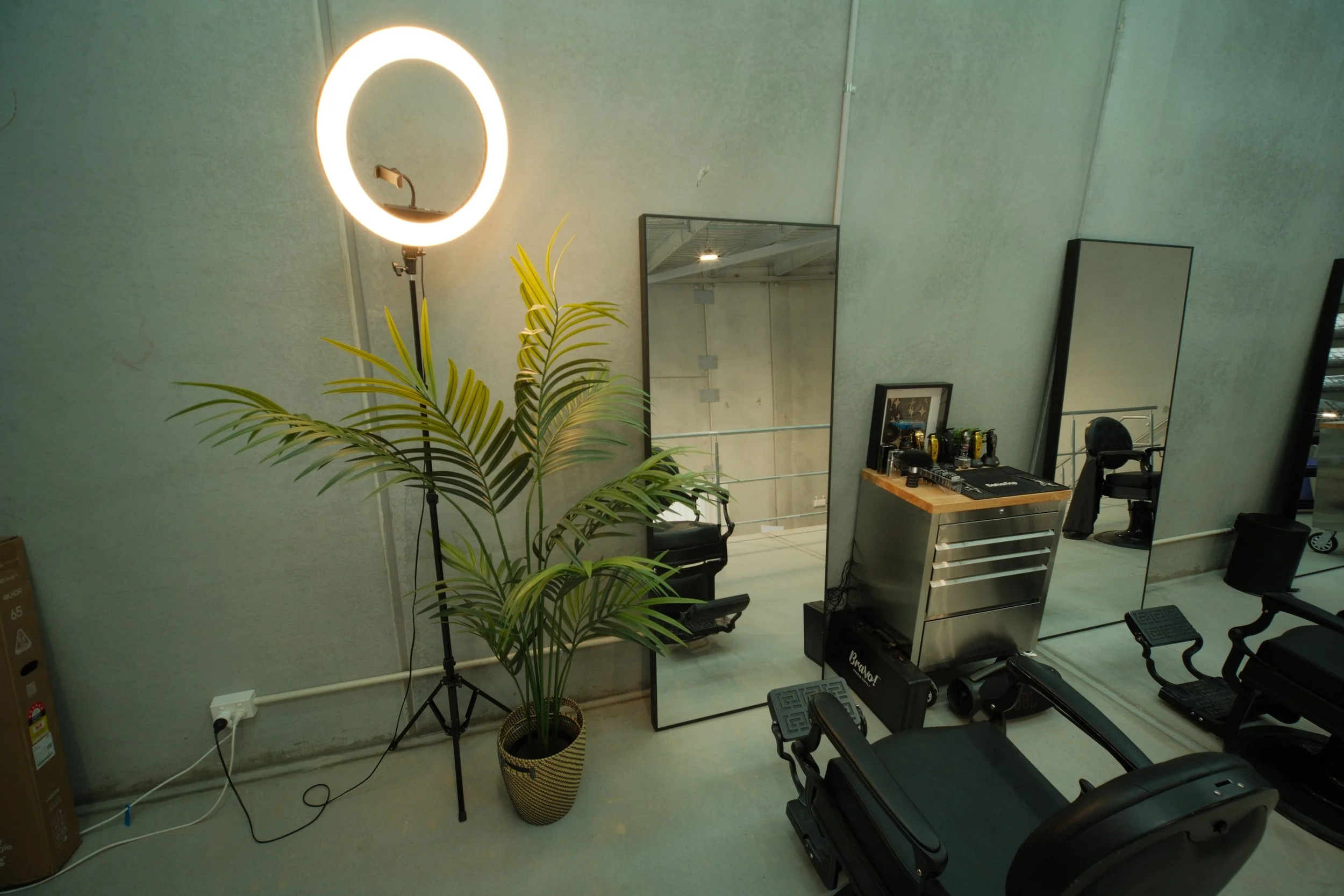 Interior view of a room with a large mirror, a ring light, a potted plant, and black office chairs. Reflection shows more mirrors, a desk with various objects, and industrial ceiling.