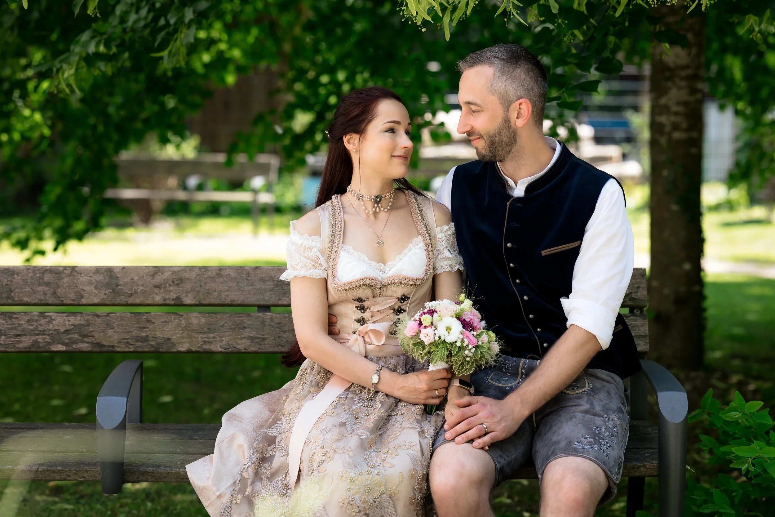 A couple dressed in traditional Bavarian clothing sitting on a park bench, with the woman holding a bouquet of flowers, looking at each other affectionately.