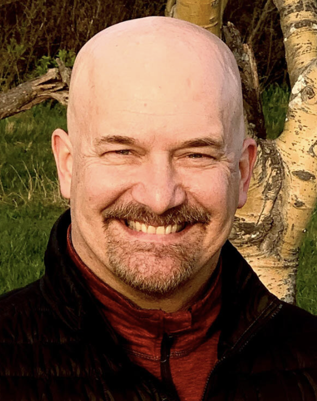 A smiling bald man with a beard and mustache, wearing a black jacket and red shirt, standing outdoors near a tree with green grass.