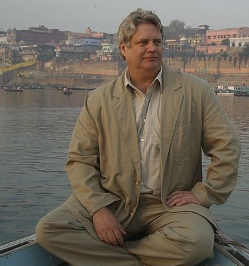 A man sitting cross-legged on a boat near a river with buildings and boats in the background.
