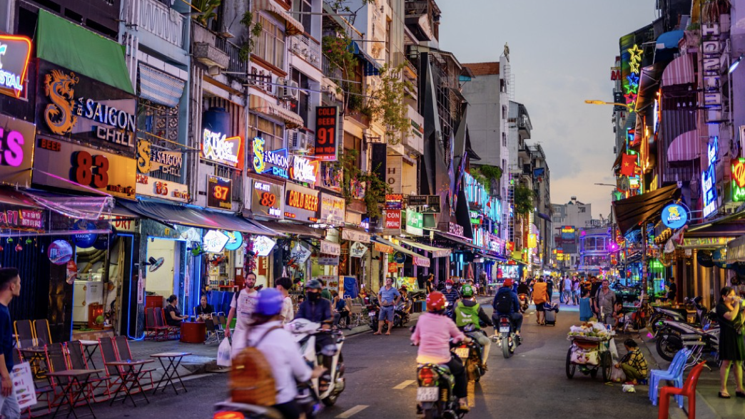 A group of people dressed in white clothing walking outdoors in a park with trees and green grass, some carrying umbrellas and bags.