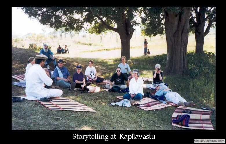 A group of people sitting on mats under a large tree, engaged in storytelling at Kapilavastu, with some participants listening and others participating, and a few people in the background.
