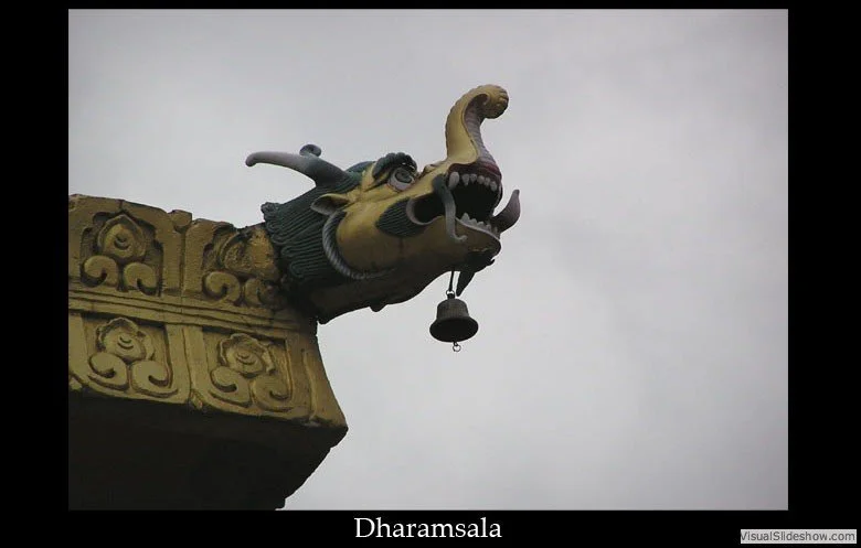 Close-up of a dragon sculpture's head with an open mouth and a hanging bell, atop a decorative structure with ornate carvings, against an overcast sky, labeled 'Dharamsala'.