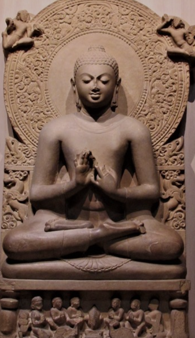 Stone sculpture of a meditative figure seated cross-legged with hands in a prayer gesture, surrounded by intricate carved patterns and mythical creatures.