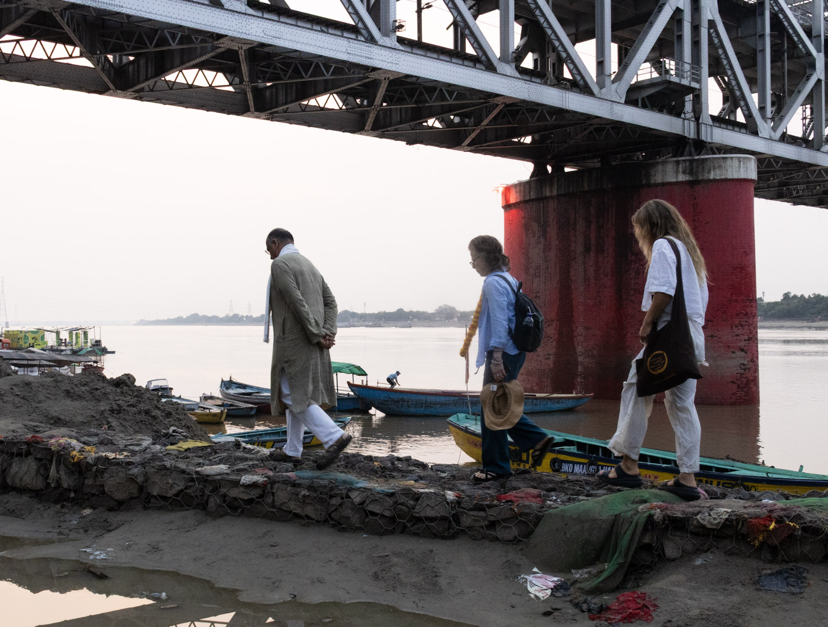 Three people walk along a riverbank under a large bridge, with boats floating nearby.