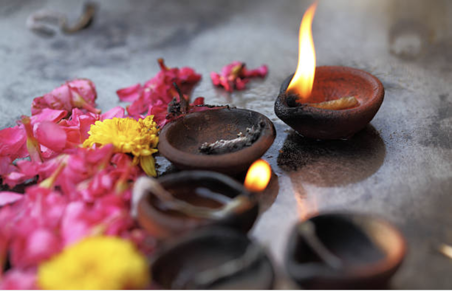 Small oil lamps with flames and scattered pink and yellow flower petals on a surface, suggesting a traditional or spiritual setting.