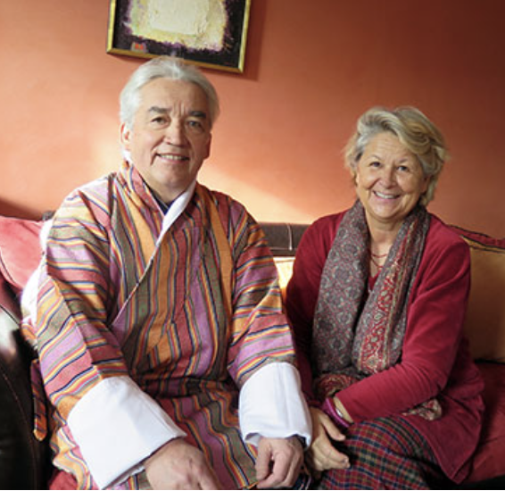 An elderly man and woman sitting on a sofa, smiling, in a cozy living room with warm-colored walls.