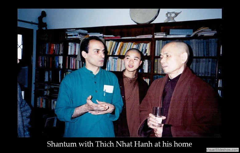 Three men are standing in a room filled with bookshelves. One man, dressed in a blue shirt, is speaking with hand gestures. The other man, in a brown jacket, is holding a glass. The third person, a young man, is listening. The room has a dark wooden 