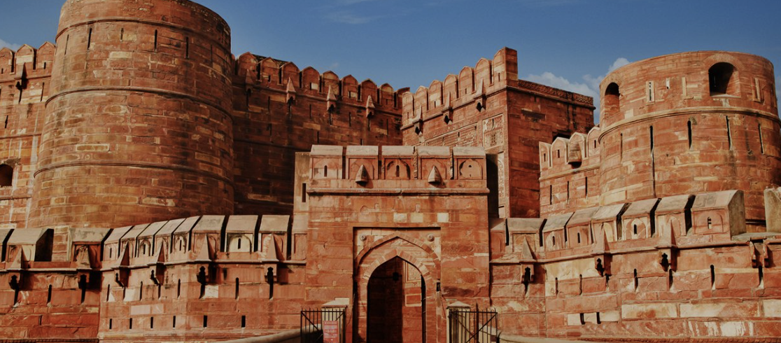The image depicts a historic red sandstone fortress with large rounded towers, a high wall, and battlements under a blue sky with some clouds.