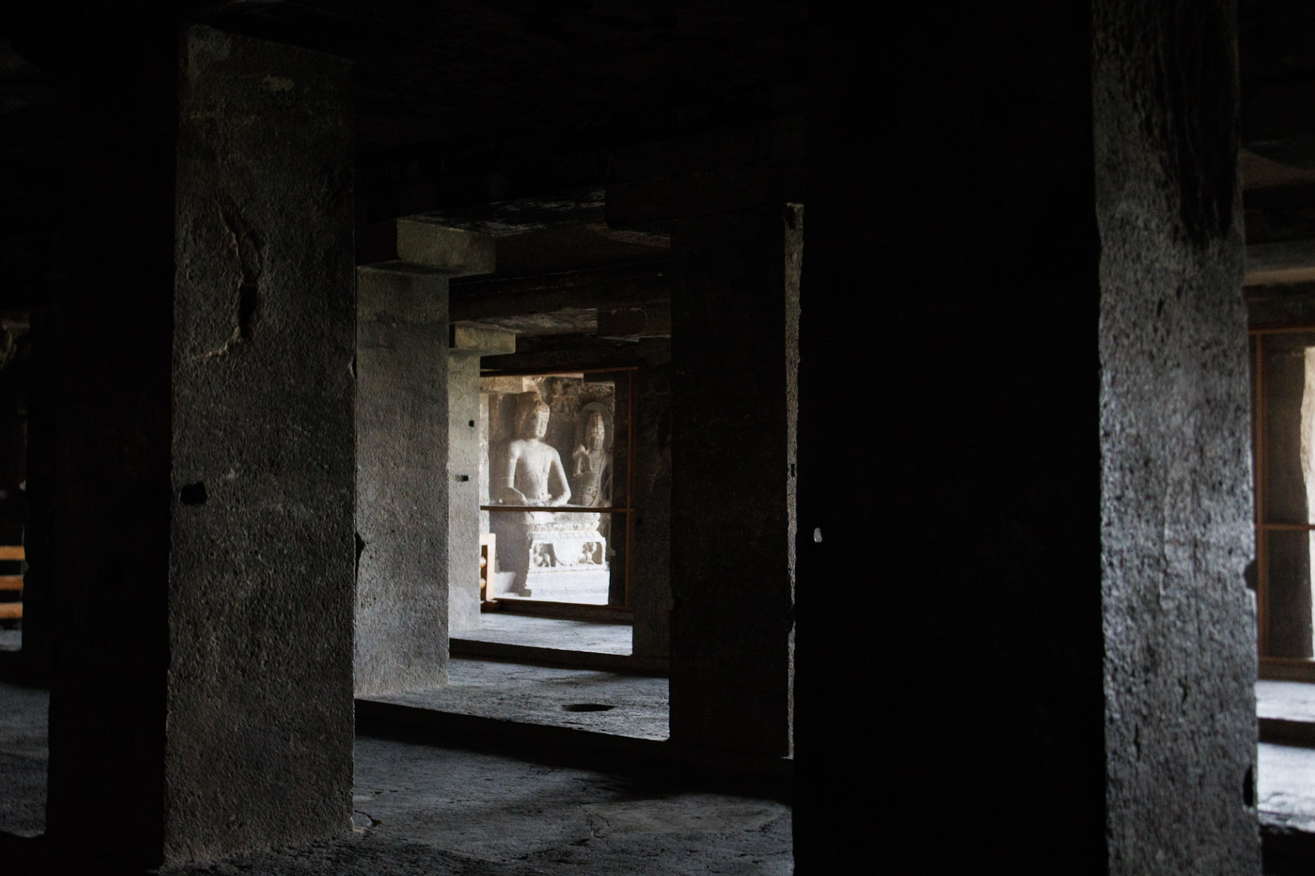 A dark interior space with stone pillars, leading to a brighter area with ancient stone sculpture of two figures in the background.
