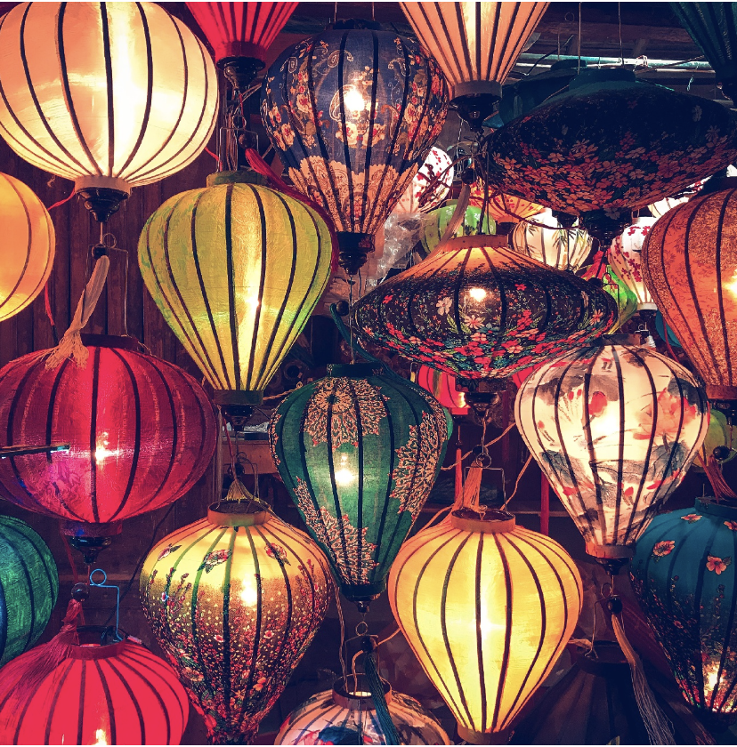 A colorful display of hanging paper lanterns in various shapes, sizes, and intricate patterns illuminating a dark space.