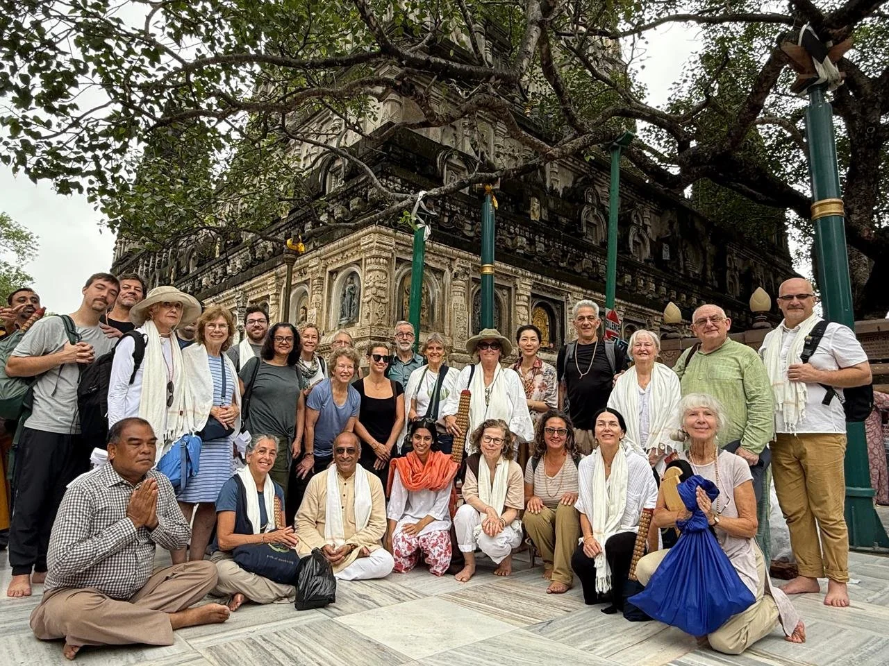 Group of people posing together in front of a historic temple or monument, some dressed in traditional attire, with a large tree and detailed stone architecture in the background.