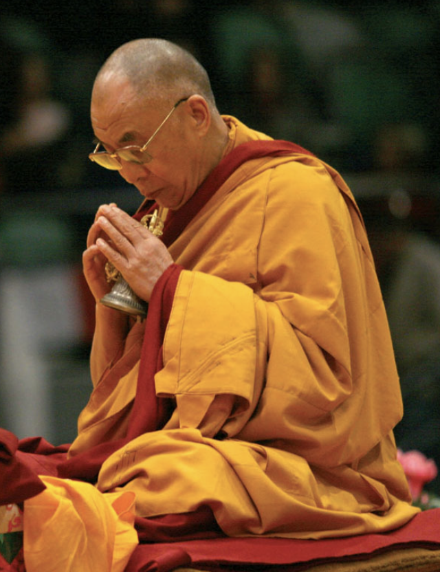 A Buddhist monk wearing orange and red robes, sitting cross-legged, holding a bell with both hands, with eyes closed in prayer or meditation.