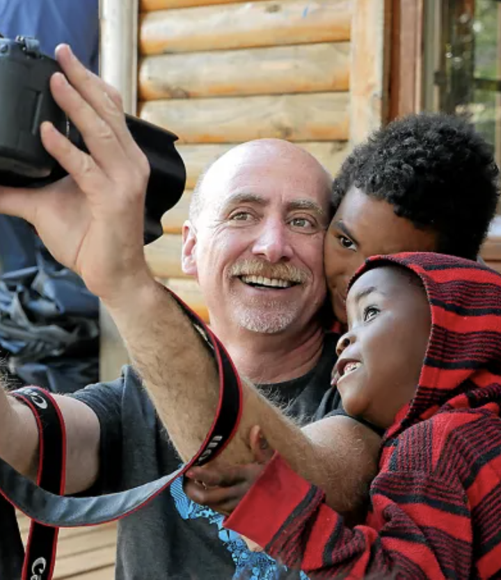 A smiling man taking a selfie with two children, one wearing a red and black hoodie, outside a wooden cabin.