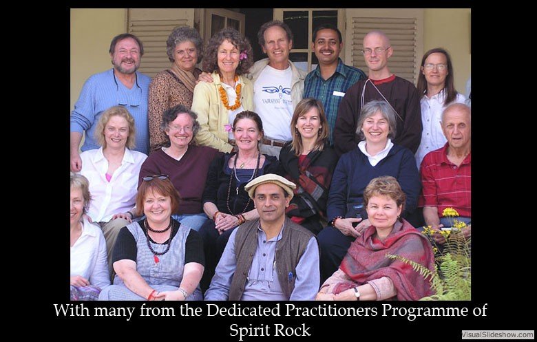 Group of people posing together outdoors with a building in the background, some wearing casual attire, others with accessories, smiling for the photo.