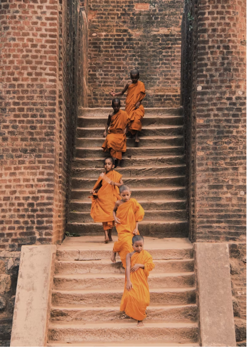 Six young boys dressed in orange robes walking down stone stairs surrounded by brick walls.