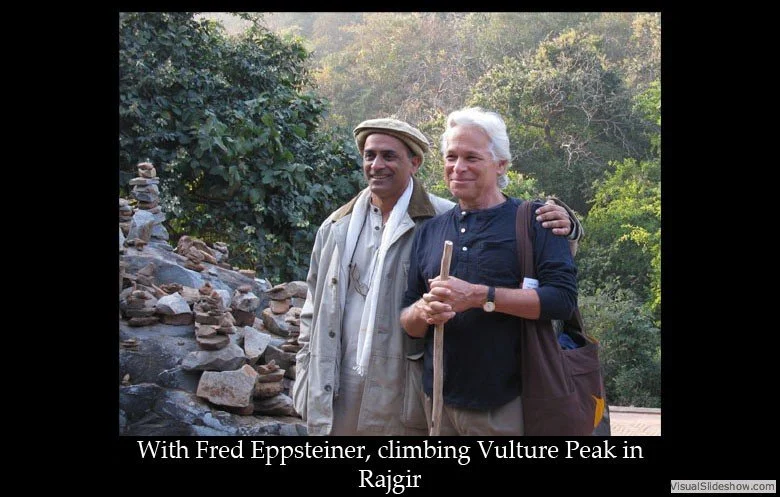 Two men posing outdoors, one with a walking stick, in front of a rocky hillside and trees, with sunlight filtering through.