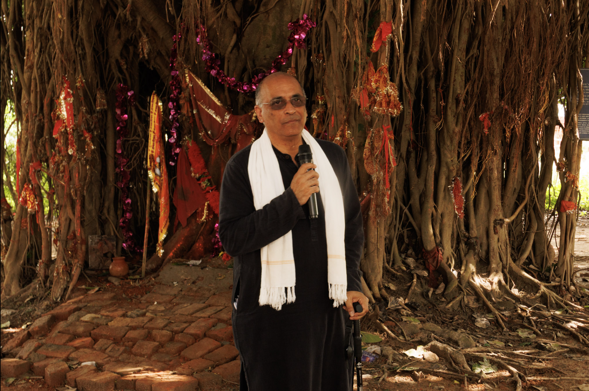 A man in a black outfit with a white scarf holding a microphone stands in front of a large tree with electrical and decorative adornments, on a brick pathway.
