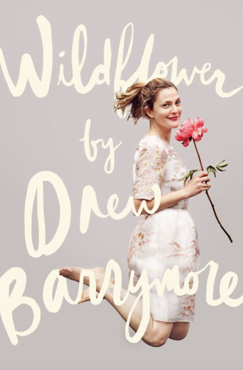 A woman with short, wavy hair holding a pink flower, kneeling with one knee on the ground, smiling, wearing a light-colored floral dress, against a plain background with the text 'Wildflower by Pre Band' overlaid.
