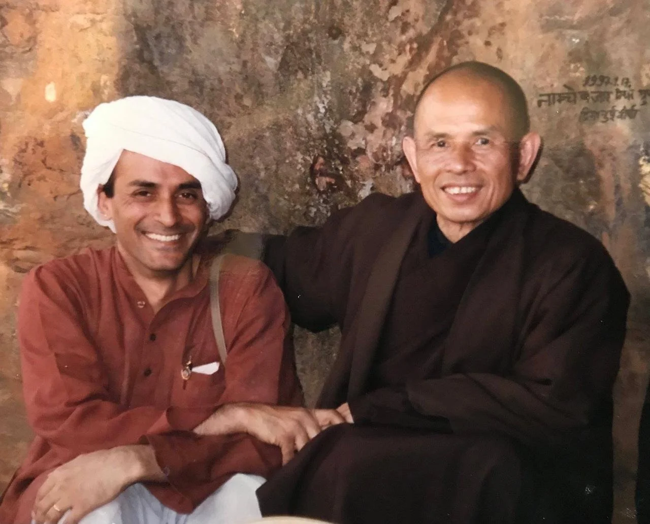 Two smiling men, one wearing a white turban and traditional Indian attire, and the other dressed in dark robes, sitting together against a textured wall background.