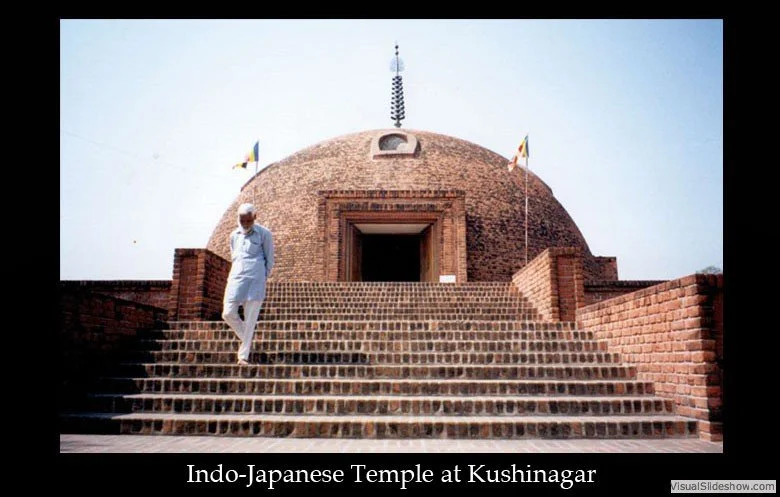 A man walking down the steps of the Indo-Japanese Temple at Kushinagar, which has a large brick-domed structure with flags on either side and a polished black border with the caption.