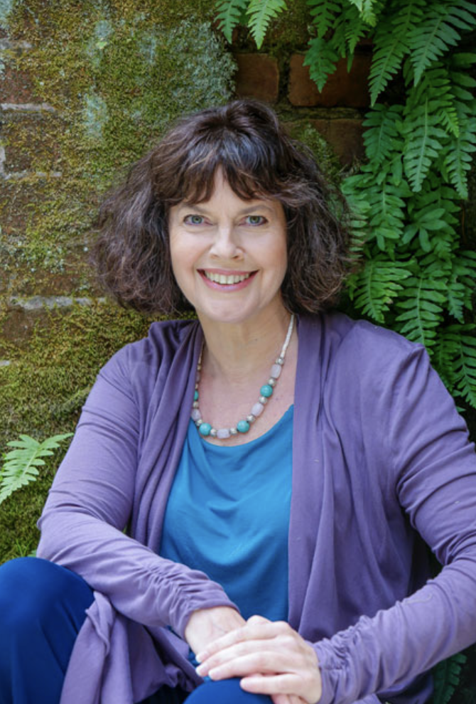 A smiling woman with dark curly hair wearing a purple jacket and blue top, sitting outdoors in front of green plants and a moss-covered brick wall.