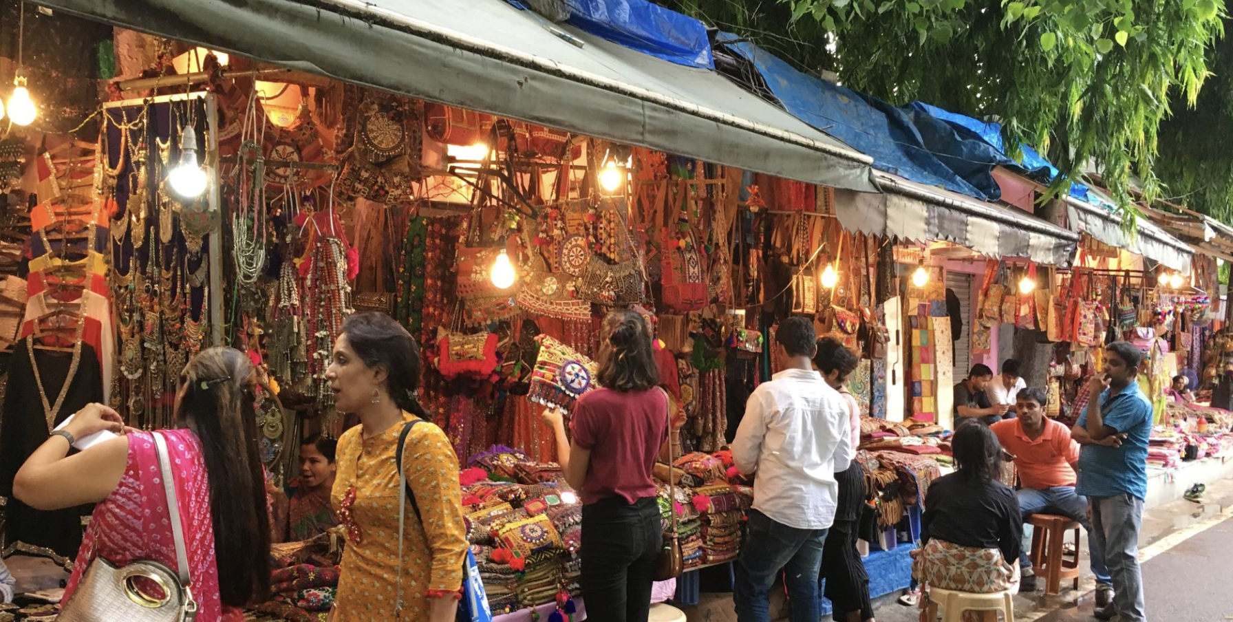 A bustling outdoor market with stalls selling colorful jewelry, bags, clothing, and accessories. Several people are walking and browsing, with some engaged in conversation, under a canopy with hanging lights. Green trees are visible overhead.