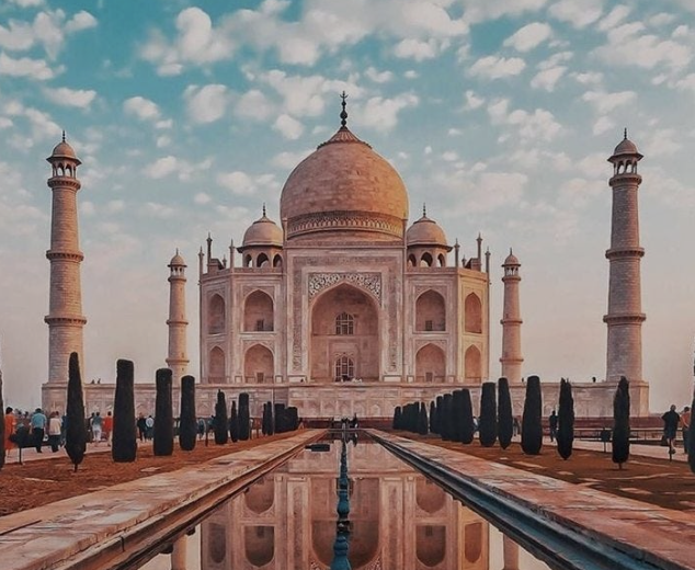 The Taj Mahal with its white marble dome and minarets, reflected in the water pool in front, under a partly cloudy sky, with visitors walking around.