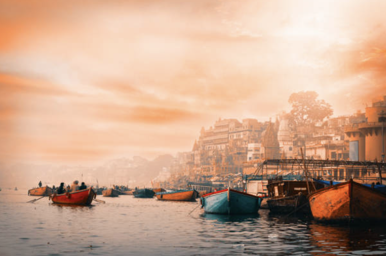 Boats floating on a river near a hillside town during sunset or sunrise with a warm, orange sky.