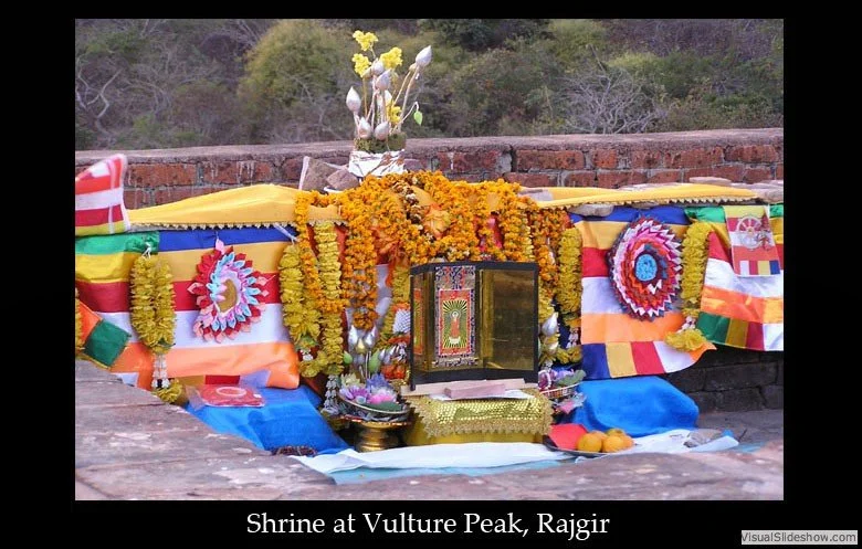 A shrine at Vulture Peak, Rajgir, decorated with marigold flower garlands, colorful cloth, and various religious items, including a framed picture and offerings, set against a stone wall with trees in the background.